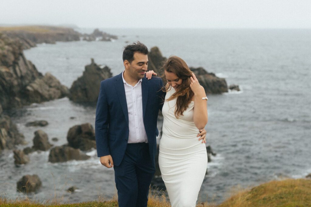 bride and groom laugh on a sea cliff with sea stacks in the background in Bonavista, Newfoundland