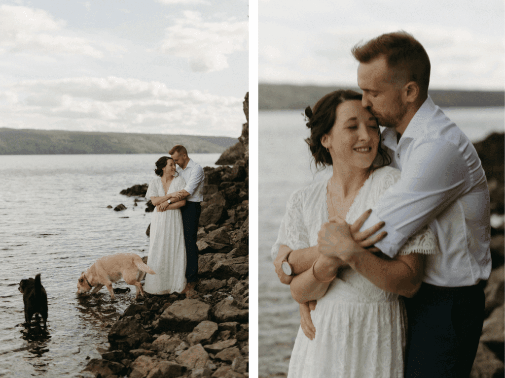 bride and groom smile calmly as they hold each other on the lake shore with their dogs playing in the water