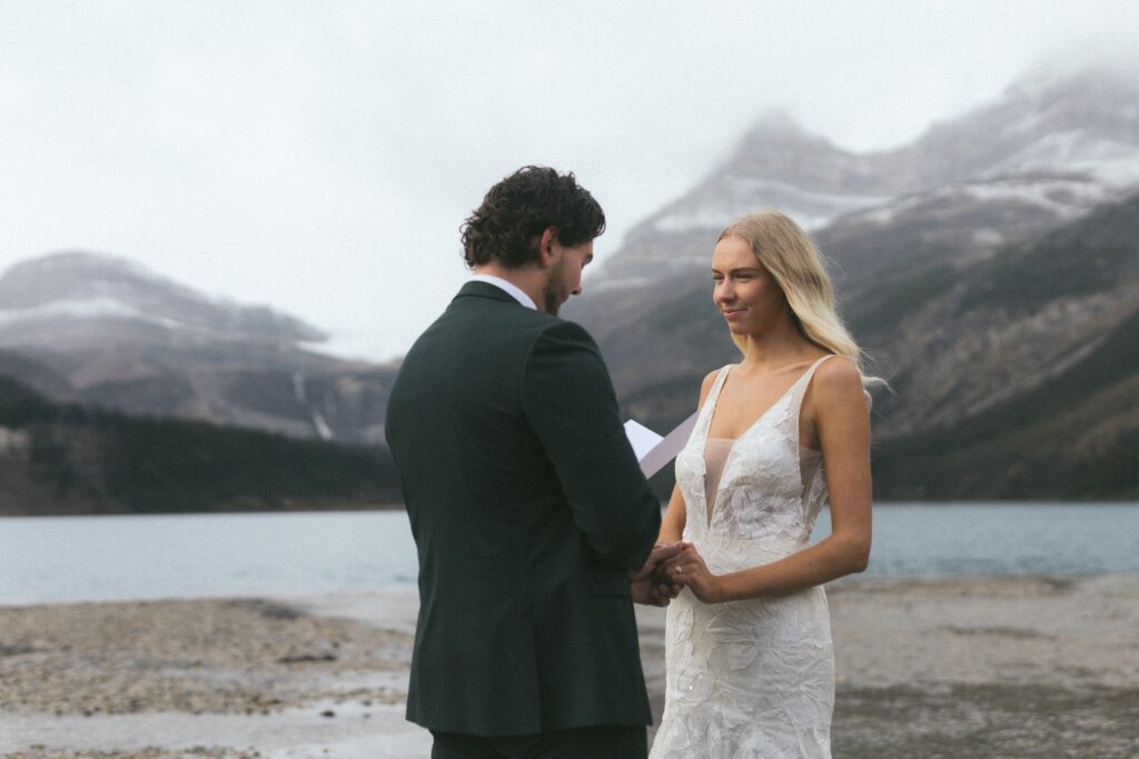 Bride smiles as groom reads vows on beach at Bow Lake in Banff with mountains rising behind