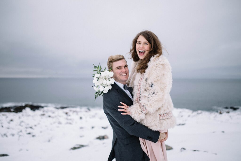 bride and groom laugh in a snowy landscape as groom picks the bride up