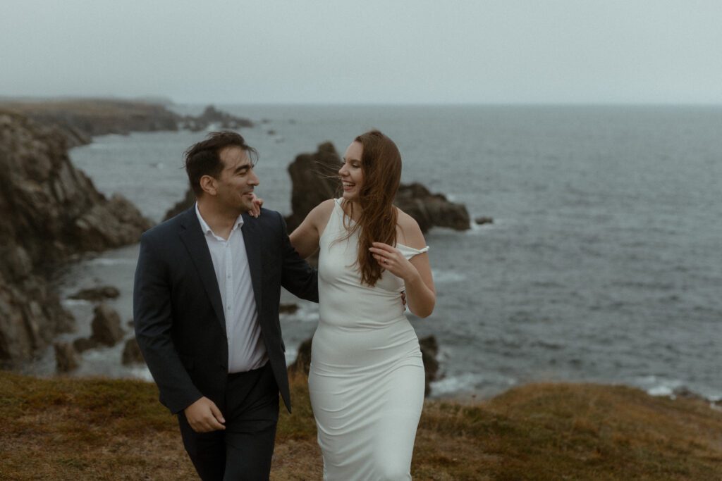 bride and groom laugh on a sea cliff with sea stacks in the background in Bonavista, Newfoundland