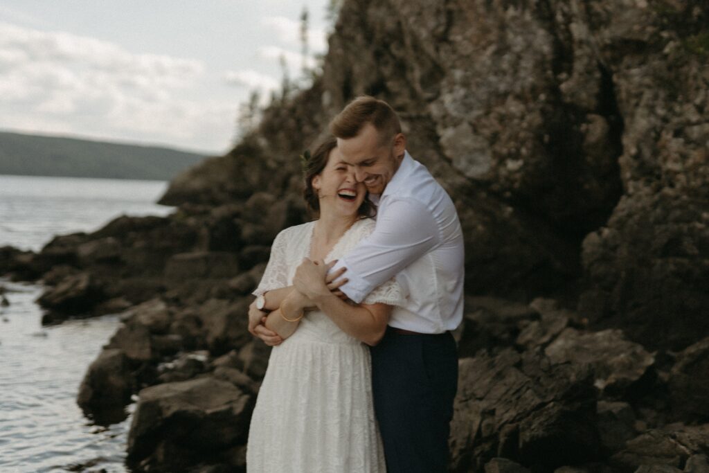 bride and groom hold each other and laugh on rocky shore