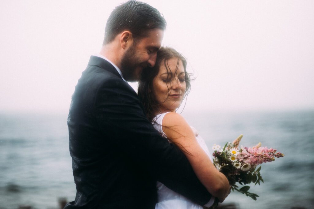 bride and groom hold each other by the sea in the pouring rain