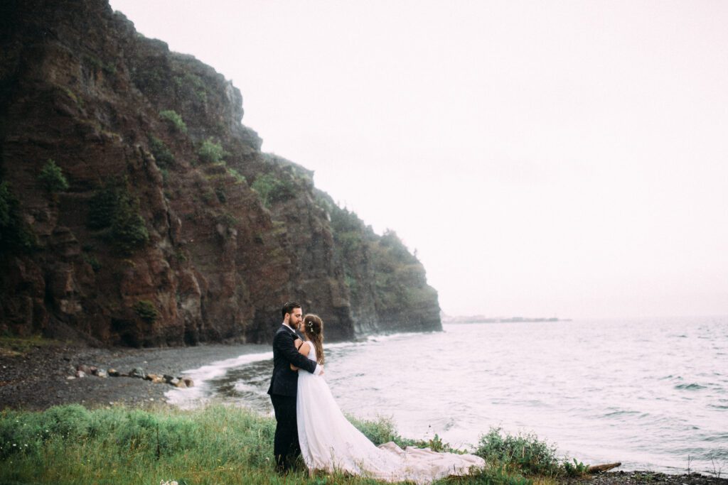 bride and groom hold each other beneath a towering sea cliff during their Newfoundland elopement