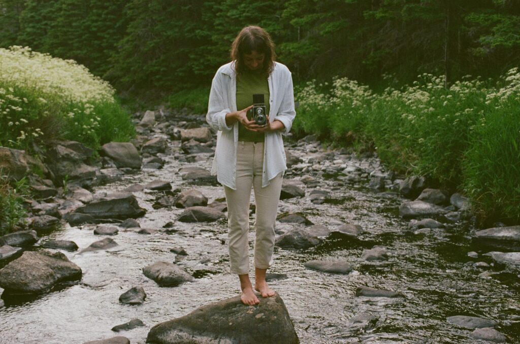 Heather stands on a rock in a river, taking a photo with a medium format film camera