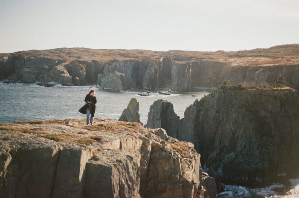 woman walks along rocky coastal ledge surrounded by sea stacks at golden hour in Spillar's Cove, Newfoundland