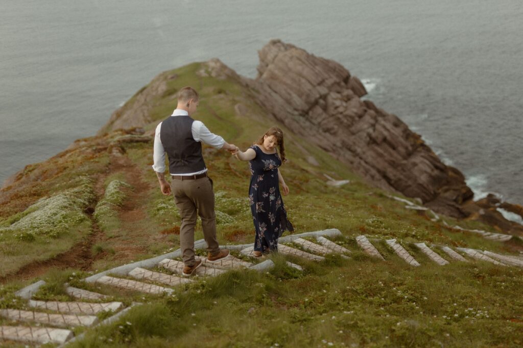 bride in a blue dress leads groom along hiking trail on a cliff above the ocean during Newfoundland elopement