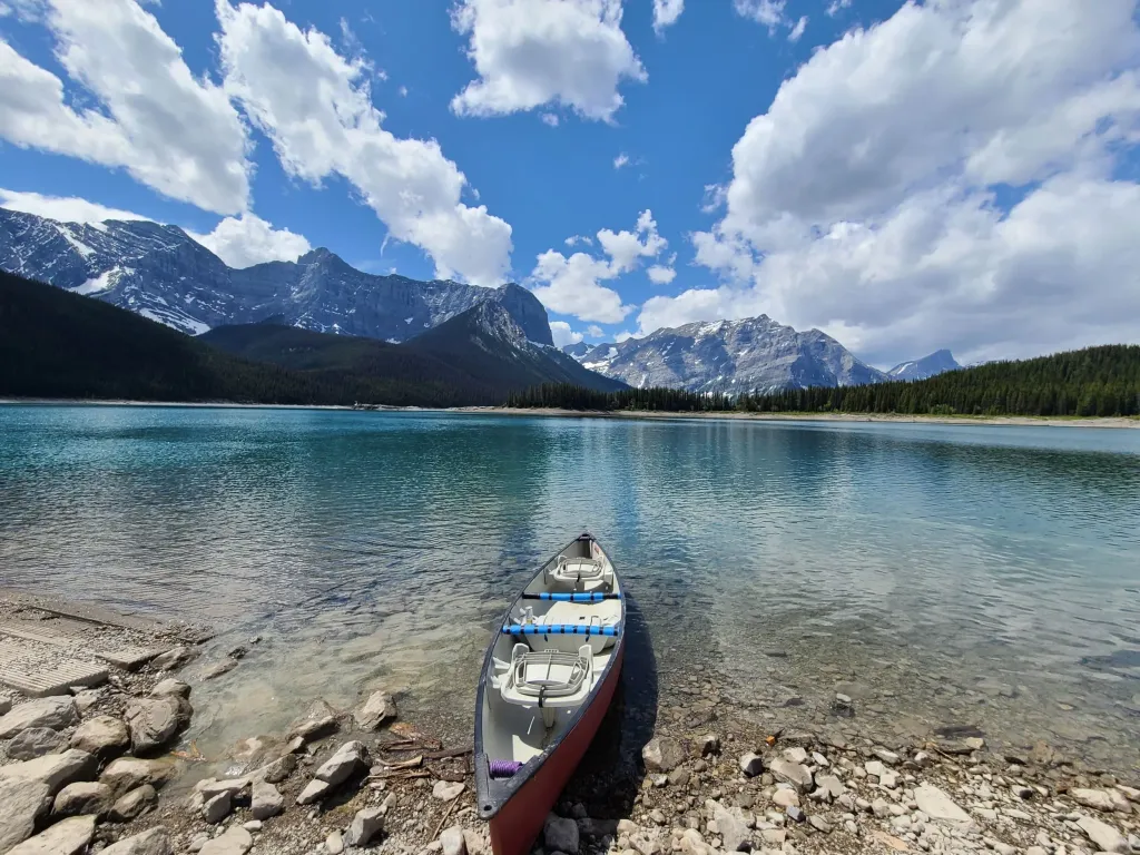 a canoe rests on the shore of a vibrant blue lake with mountains rising in the background in Kananaskis, Alberta