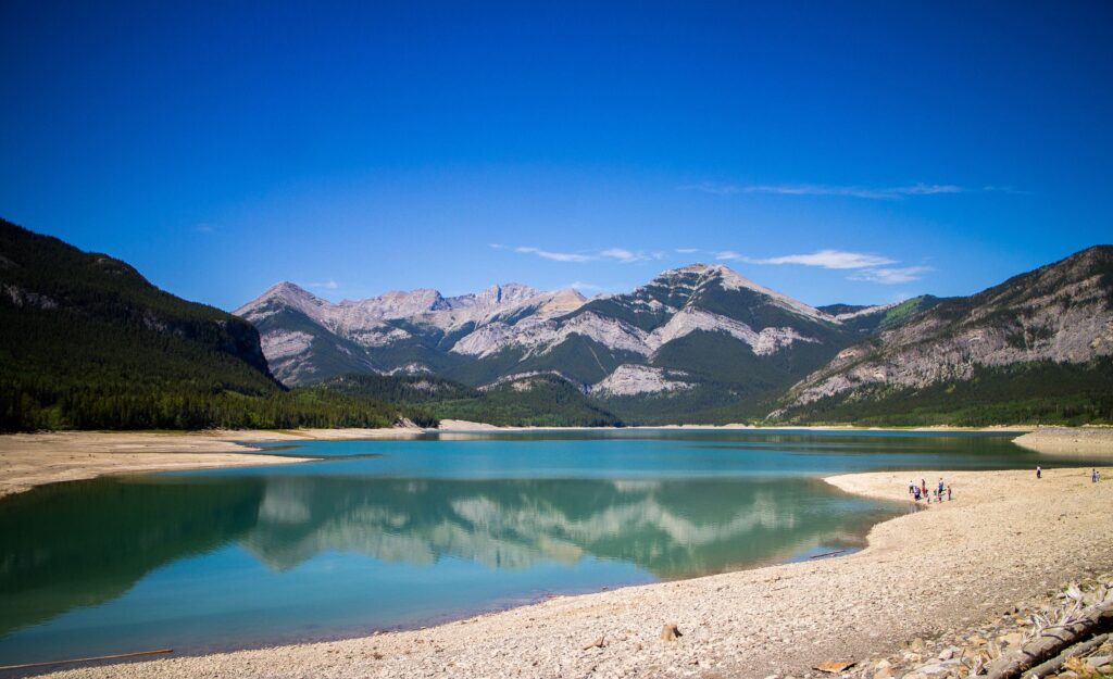 a sunny day on Barrier Lake, Kananaskis with a clear blue lake, distant people playing on the beach, and mountains rising in the background.
