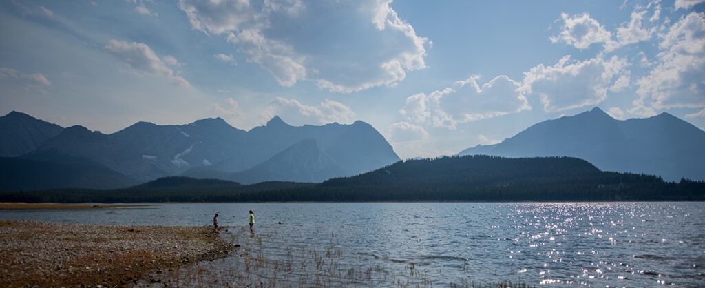 two people wade in to Lower Kananaskis Lake with mountains in the background
