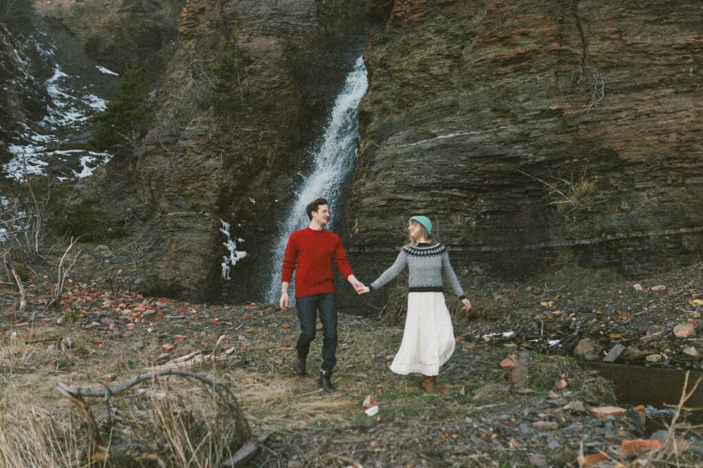 couple walk past waterfall and towering cliffs