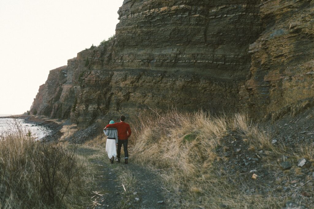 couple in wool sweaters walk along the base of tall cliffs