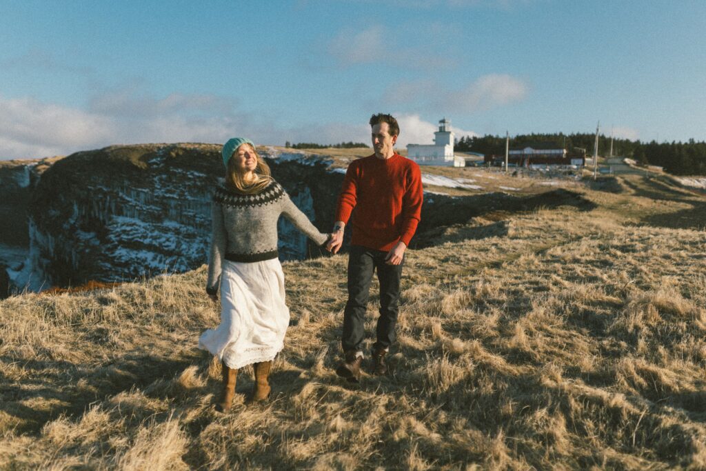 couple in wool sweaters walk in front of a lighthouse