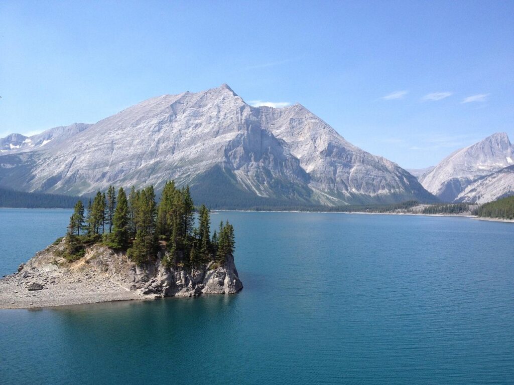 Upper Kananaskis lake on a sunny day, with a small peninsula in the foreground and mountains rising in the background