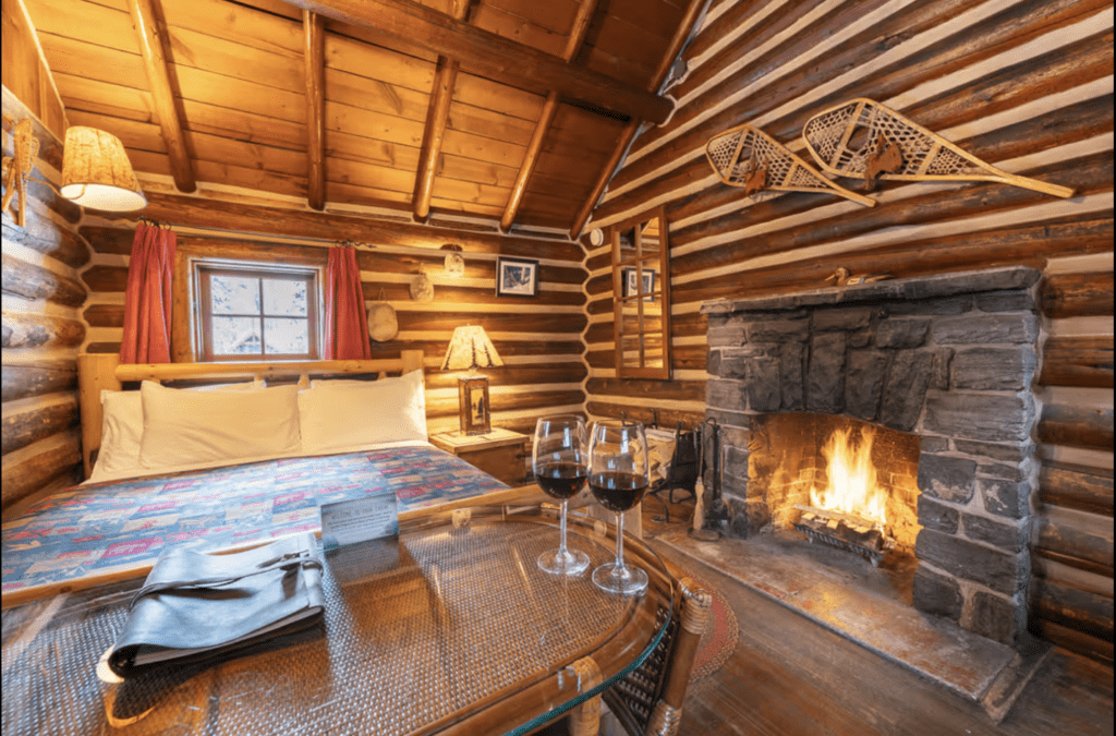 the interior of a small log cabin with a fire burning in a stone fireplace and snowshoes hanging above it.