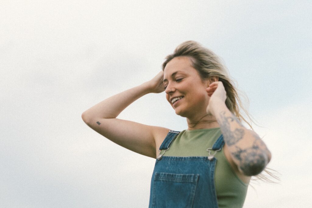 blonde woman with tattoos laughs with a cloudy sky in the background