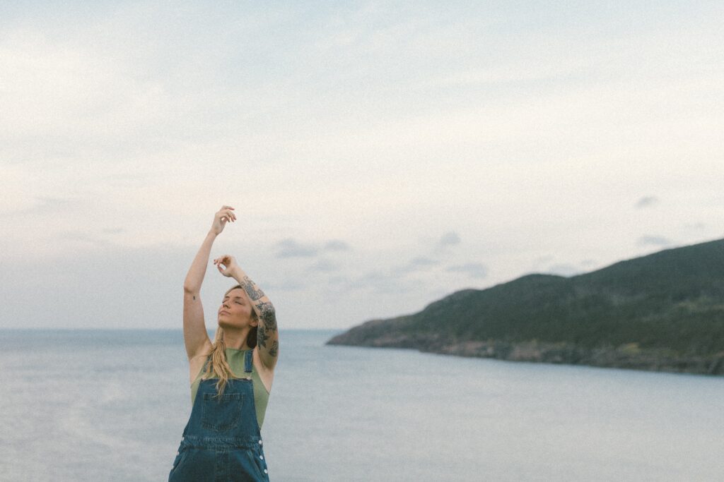 blonde woman with tattoos dances with her arms up and eyes closed by the ocean during Self Love Session