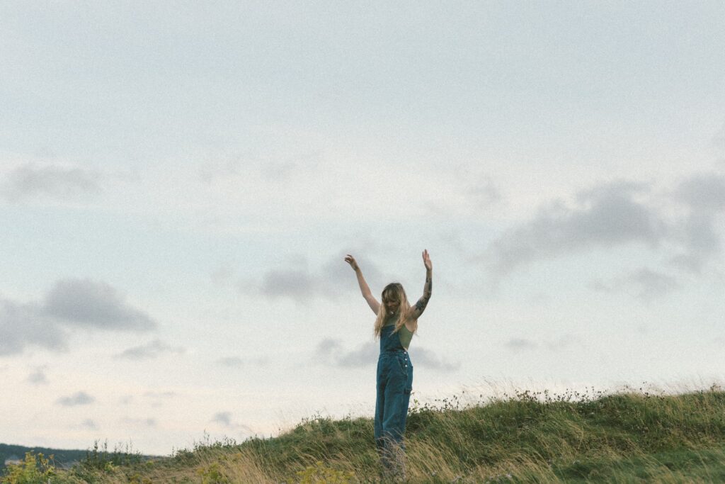 blonde woman with tattoos dances in a grassy meadow as the sun sets