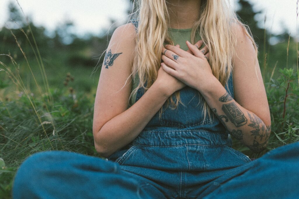 white woman with tattoos holds her hands over her heart while sitting cross legged in a meadow