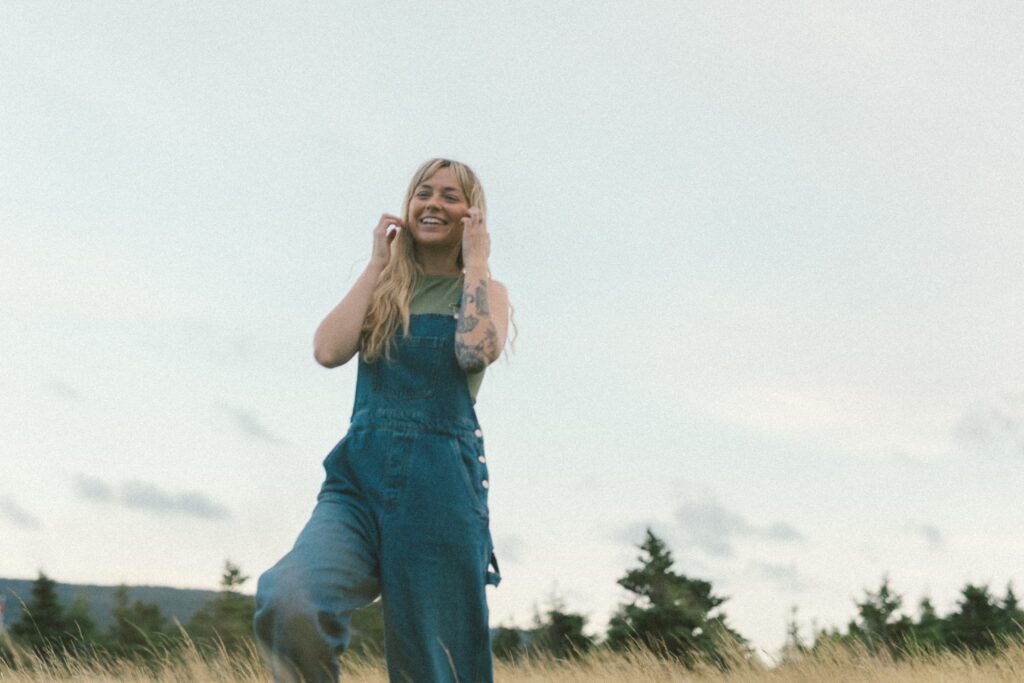 blonde woman with tattoos laughs in a field of tall grass at sunset