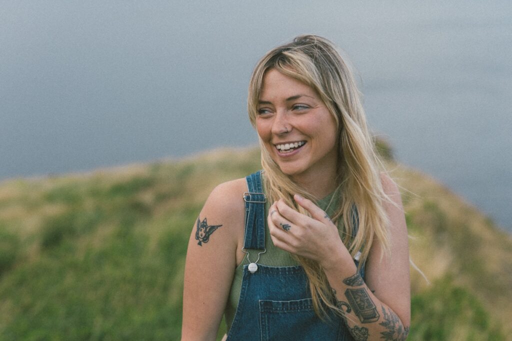 blonde woman with tattoos laughs in a grassy meadow by the ocean