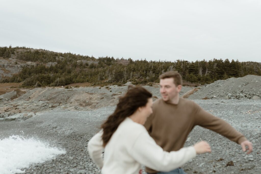 candid photo of a couple in hiking attire running from the waves on a rocky beach