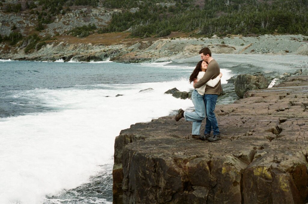film photo of a couple hugging on a rocky outcrop above the waves in Newfoundland