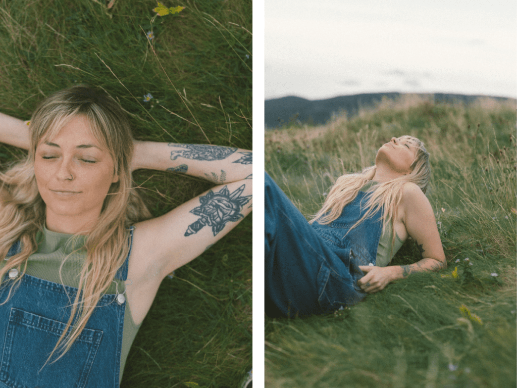 blonde woman with tattoos lays in the tall grass with her eyes closed and a peaceful smile during Self Love Session
