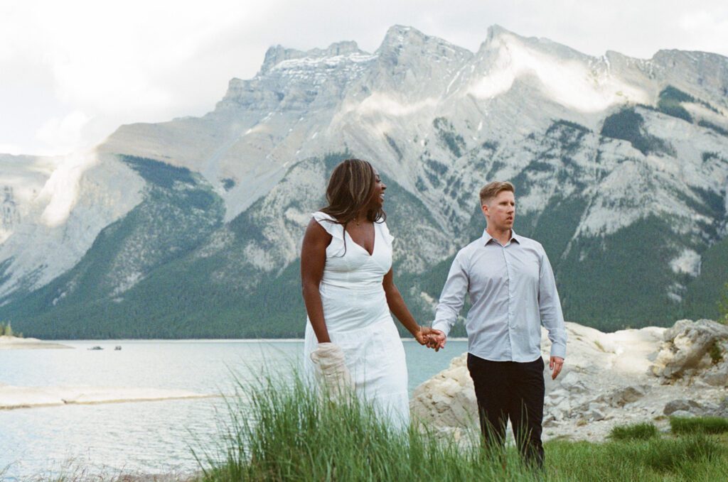 bride and groom hold hands as they wander along the shore at a mountain lake in Banff