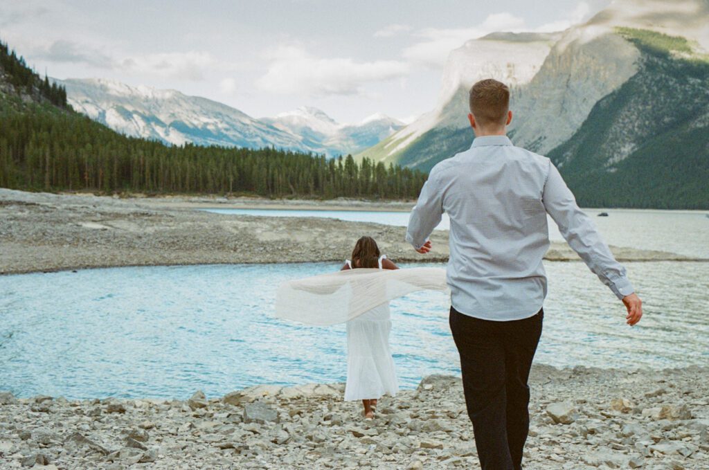 Groom walks towards bride, who is twirling on the shore of Lake Minnewanka arrounded by mountains.