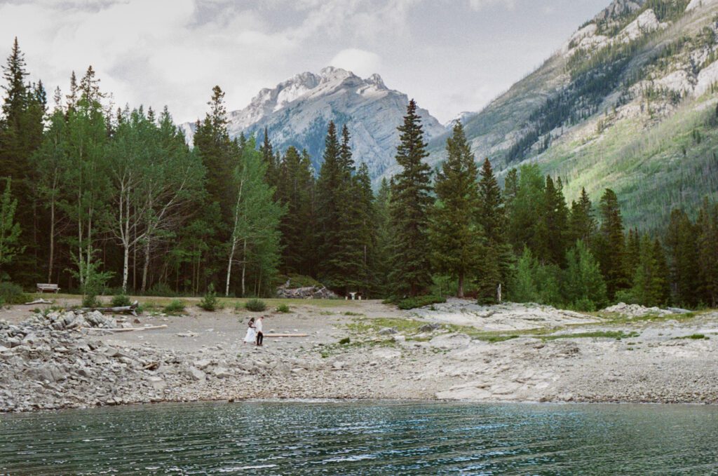 bride and groom wander the shoreline of Lake Minnewanka in Banff with mountains in the background