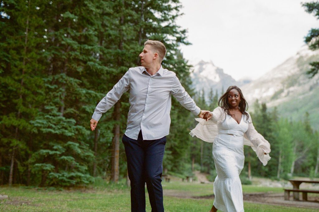 bride and groom hold hands and spin around in a lush green campground in Banff National Park