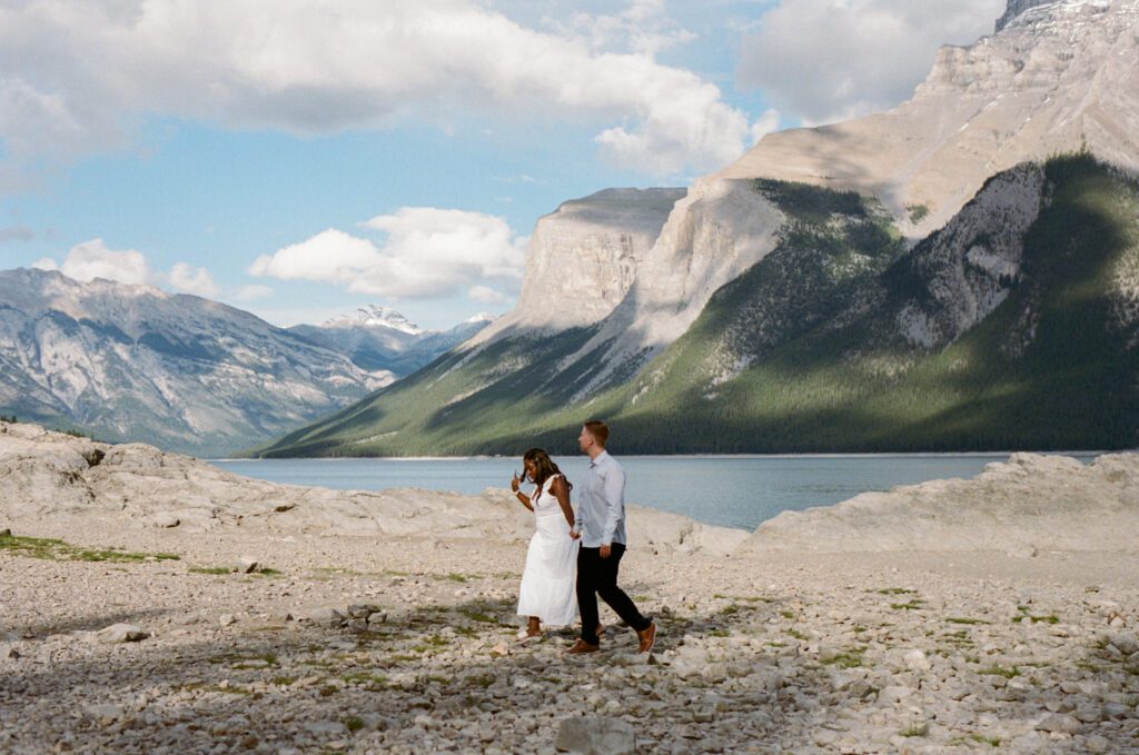 bride and groom walk along the rocky shoreline of Lake Minnewanka in Banff with mountains in the background.