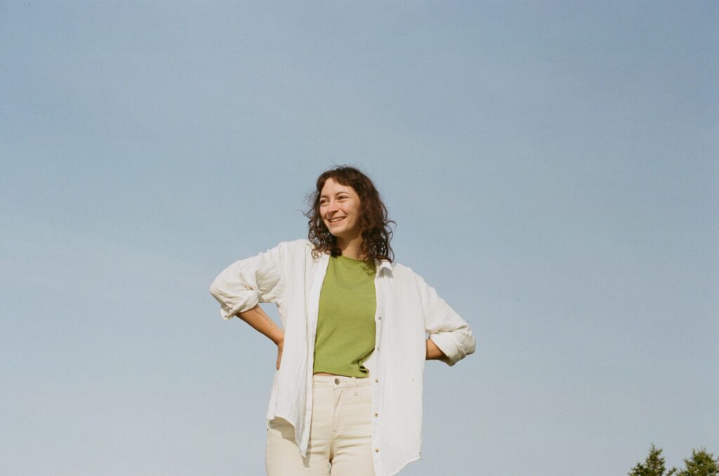 Heather laughs with their hands on their hips and the blue sky behind them.