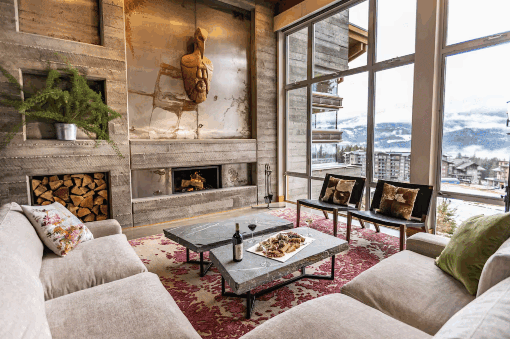 a spacious interior with cement walls, warm lighting, and modern furnishings looks out over a ski town with snowy mountains