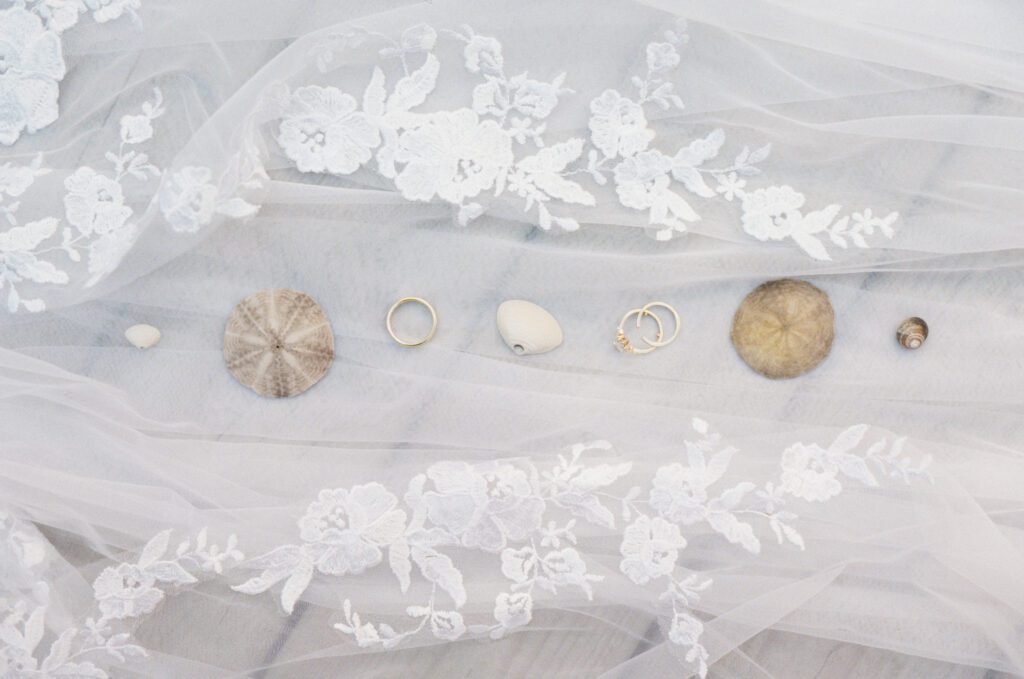 wedding rings and seashells are laid out in a neat line along the lacy edge of a veil