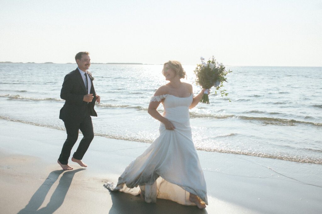 bride and groom run through the wet sand on the beach at sunset