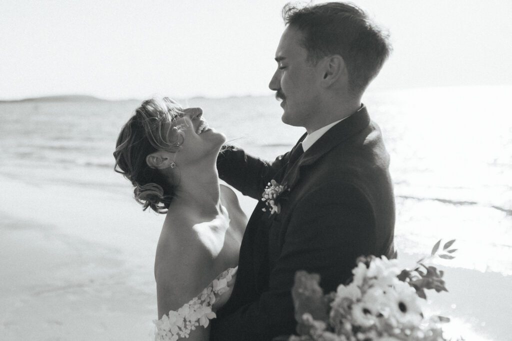 bride and groom laugh and hold each other close on the beach at sunset