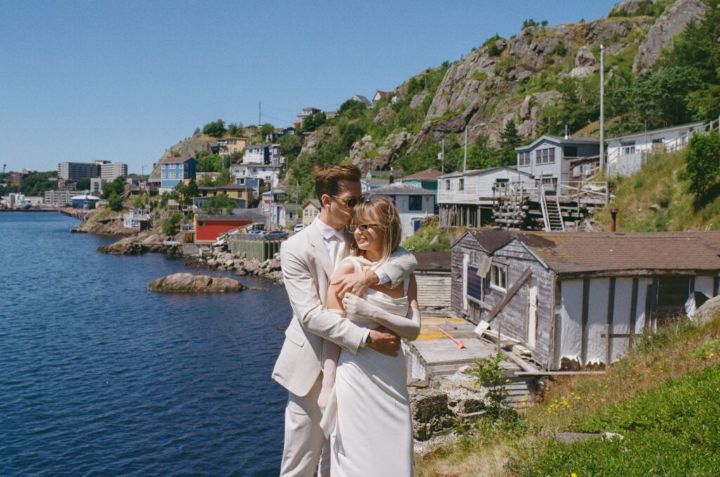 35 mm photo of bride and groom hugging in The Battery neighbourhood of St. John's with fishing shacks behind them