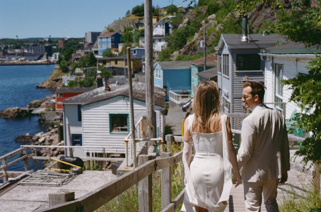 film photo of bride and groom walking throughThe Battery neighbourhood of St. John's with fishing shacks in background
