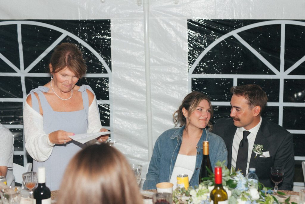 bride and groom smile at each other as bride's mother reads speech