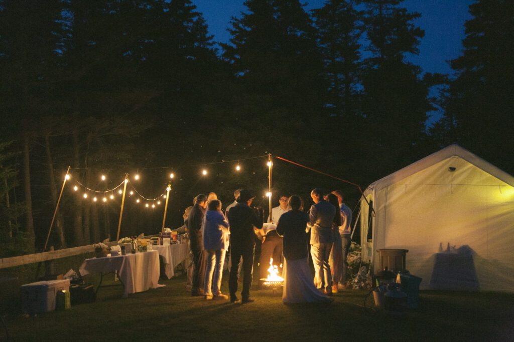 wedding guests stand in a circle around the campfire with twinkle lights all around them