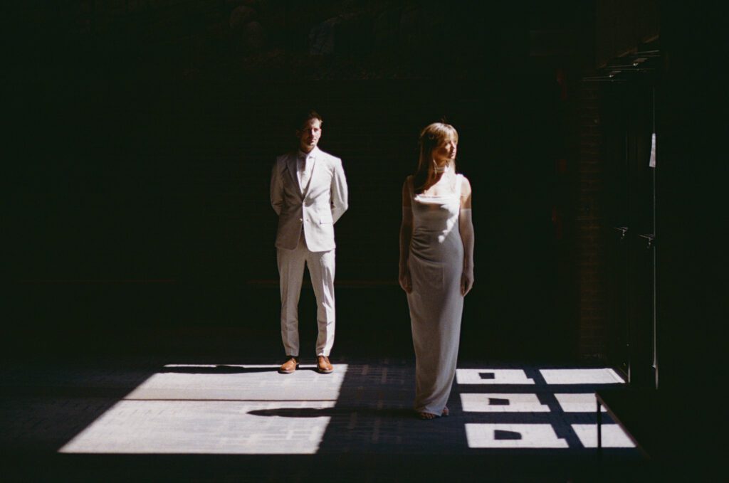 bride and groom stand in window light while the rest of the room is in shadow