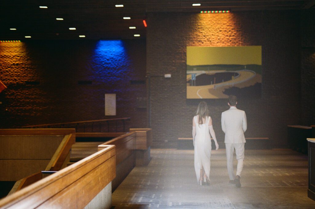 35 mm photo of bride and groom walking toward a large yellow painting in a brick building