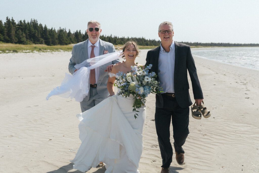 a bride with a blue bouquet walks up the beach with two fathers