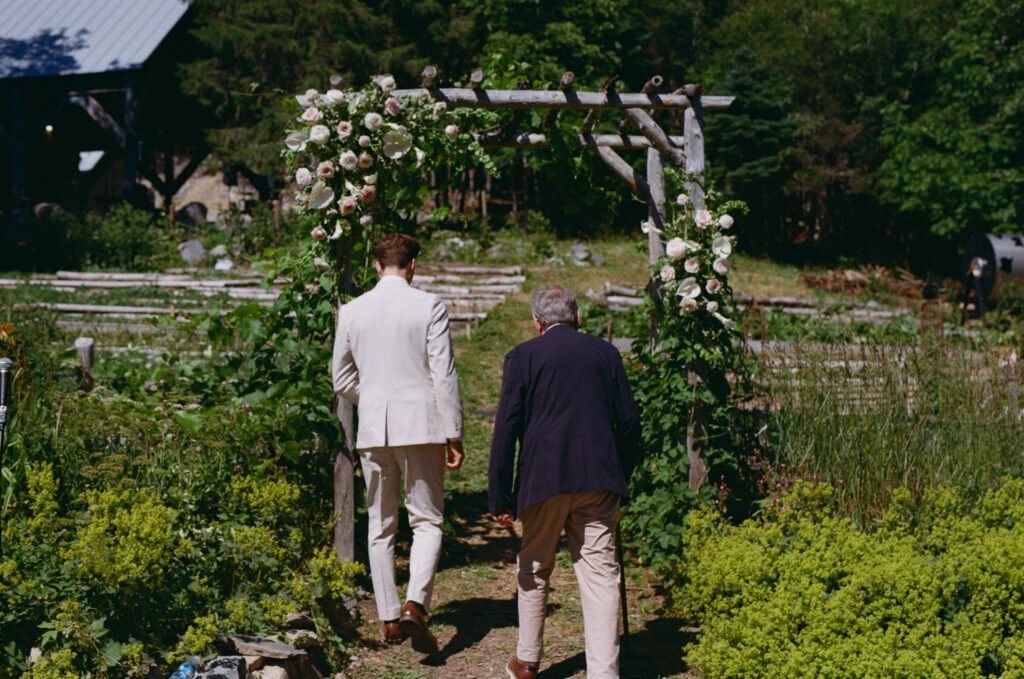 35 mm photo of groom and father walking to the ceremony arch decorated with flowers with garden beds in background