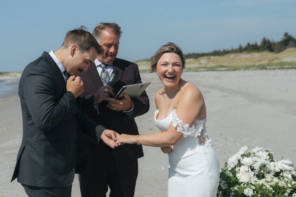 bride laughs while the groom puts ring on her finger during beach wedding in Newfoundland