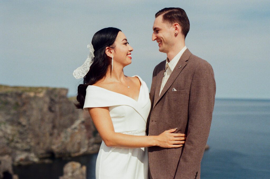 bride and groom smile at each other beside the ocean and rocky cliffs