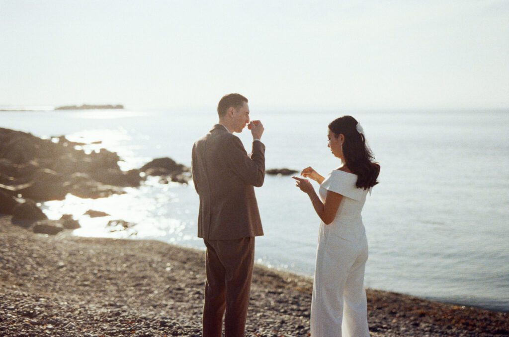 bride and groom eat ice cream on a rocky beach with sun glinting on the water