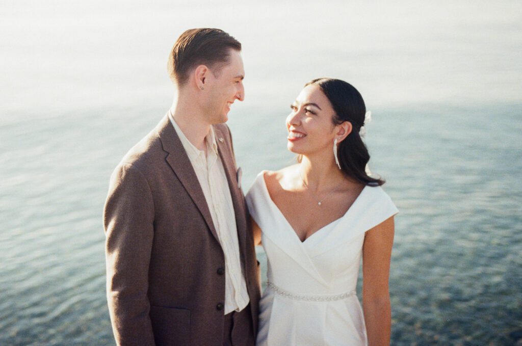 bride and groom smile at each other beside the ocean